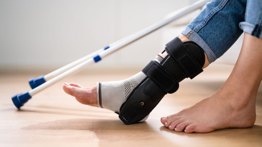 A person with a foot cast and medical boot sits on a wooden floor, crutches beside them. The scene conveys a sense of recovery and healing.