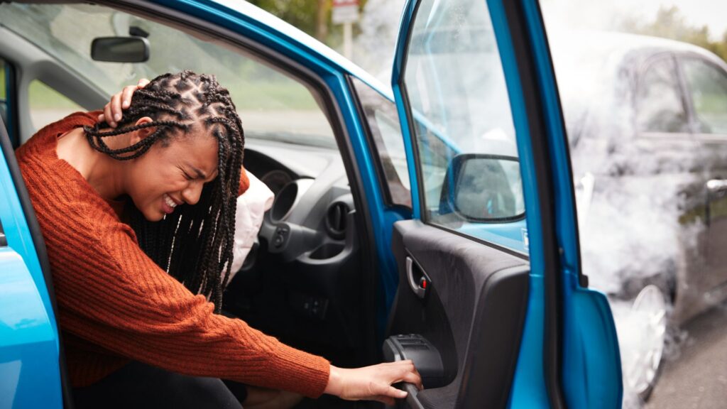A woman in a blue car holds her head in pain, appearing stressed. Smoke billows from a nearby car, suggesting a recent accident. She wears a concerned expression.