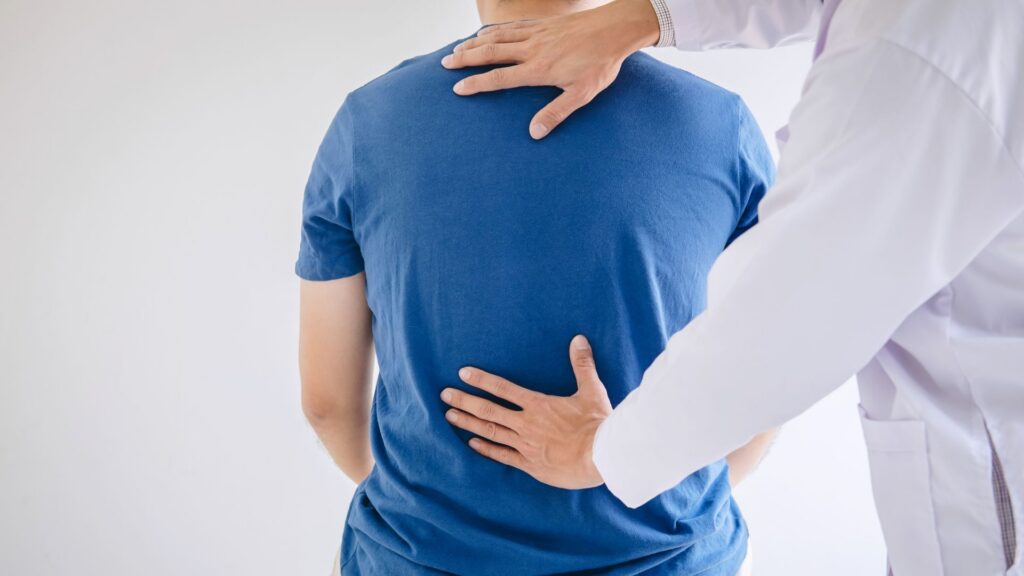 A doctor assesses a patient's back, placing hands on upper and lower spine. The patient wears a blue shirt. The setting is clinical, suggesting care.