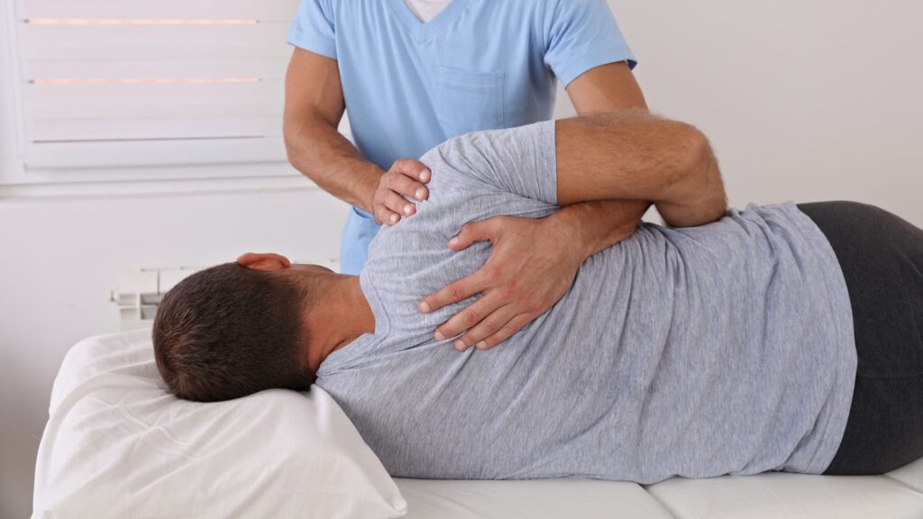 A physical therapist in a blue uniform adjusts a man's back as he lies on his side on a treatment table, conveying a sense of care and support.