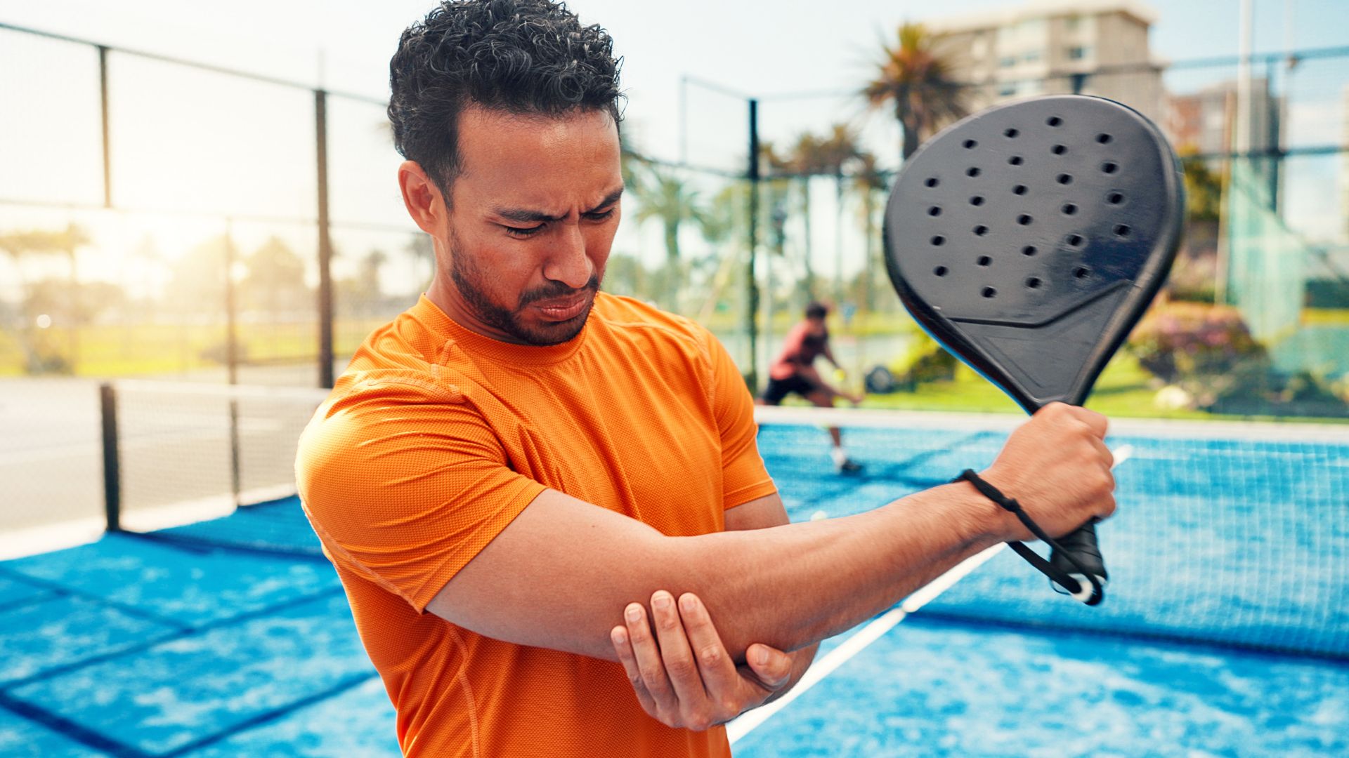 Man in an orange shirt holds his elbow in pain on a paddle tennis court, conveying discomfort. Another player is visible in the blurred background.