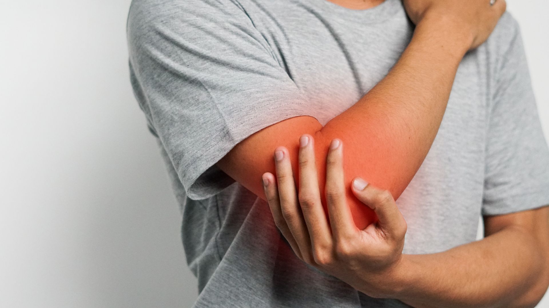 A person in a gray shirt is holding their red, inflamed elbow, suggesting pain or discomfort. The tone conveys concern and physical discomfort.