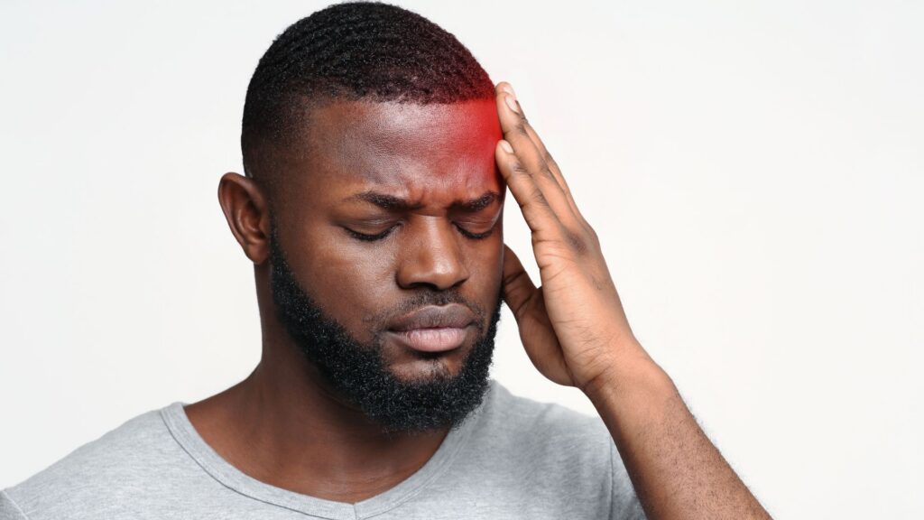A man with closed eyes holds his head, wincing in pain, with a red glow on his forehead indicating a headache. He wears a gray shirt against a white background.