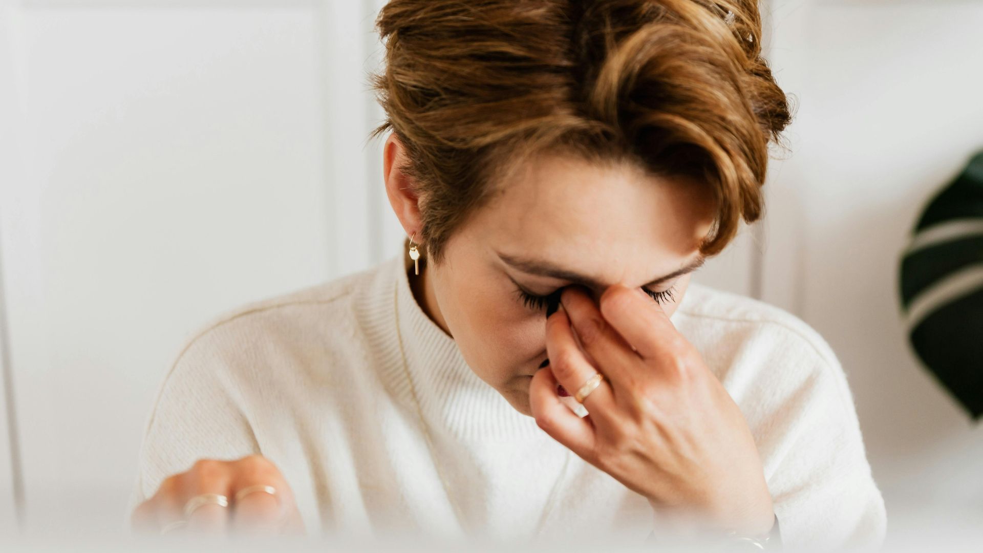 Woman in a white sweater rubbing her eyes with a hand, conveying fatigue or stress. Neutral background suggests an indoor setting.