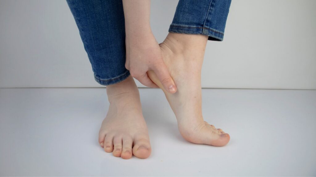 A person wearing blue jeans holds their right foot, seemingly in discomfort or pain, against a plain white background.