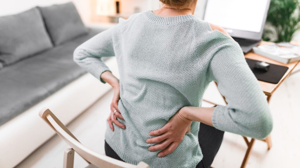 A person in a light sweater sits on a chair with hands on their lower back, indicating pain. A desk, computer, and sofa are in the bright room.