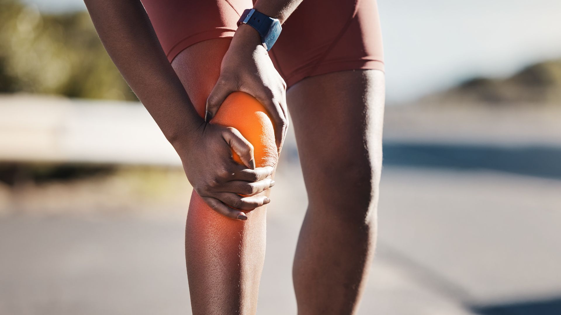 A person in athletic shorts holds their knee in discomfort, highlighted with an orange glow, suggesting pain. Outdoor setting, displaying a fitness scenario.
