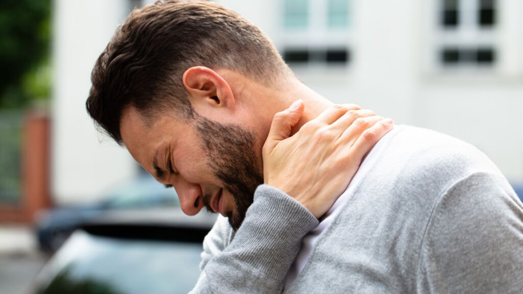 A man in a gray sweater clutches his neck, wincing in pain outdoors. The background is blurred, conveying discomfort or strain.
