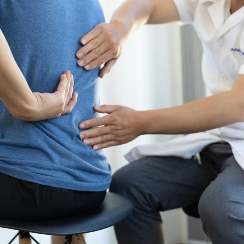 A healthcare professional gently examines a person's lower back, placing hands for assessment. The person sits on a stool, wearing a blue shirt and black pants.