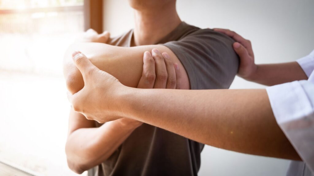 A therapist in a white coat assists a person in a gray shirt with an arm stretch in a bright room, conveying a sense of care and rehabilitation.