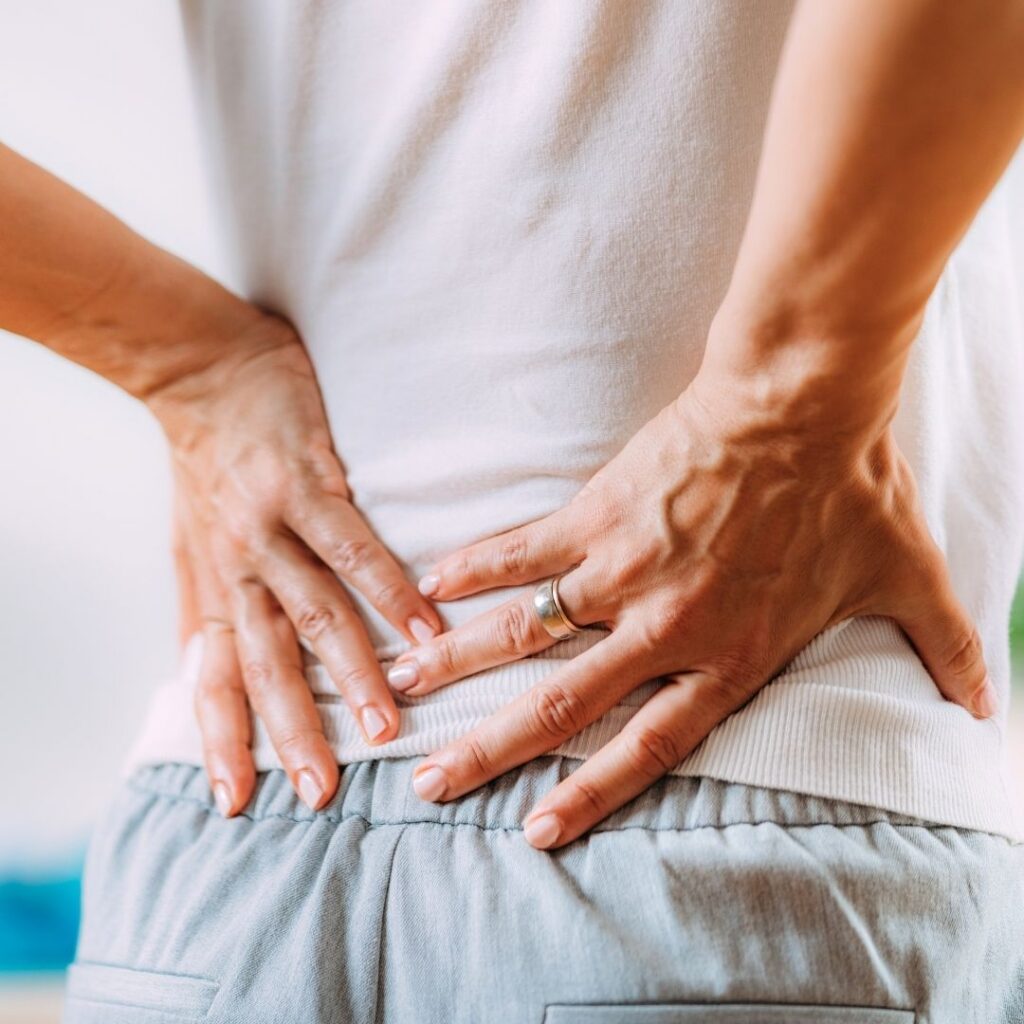 Close-up of a person wearing a white shirt and gray pants, holding their lower back with both hands, suggesting discomfort or back pain.