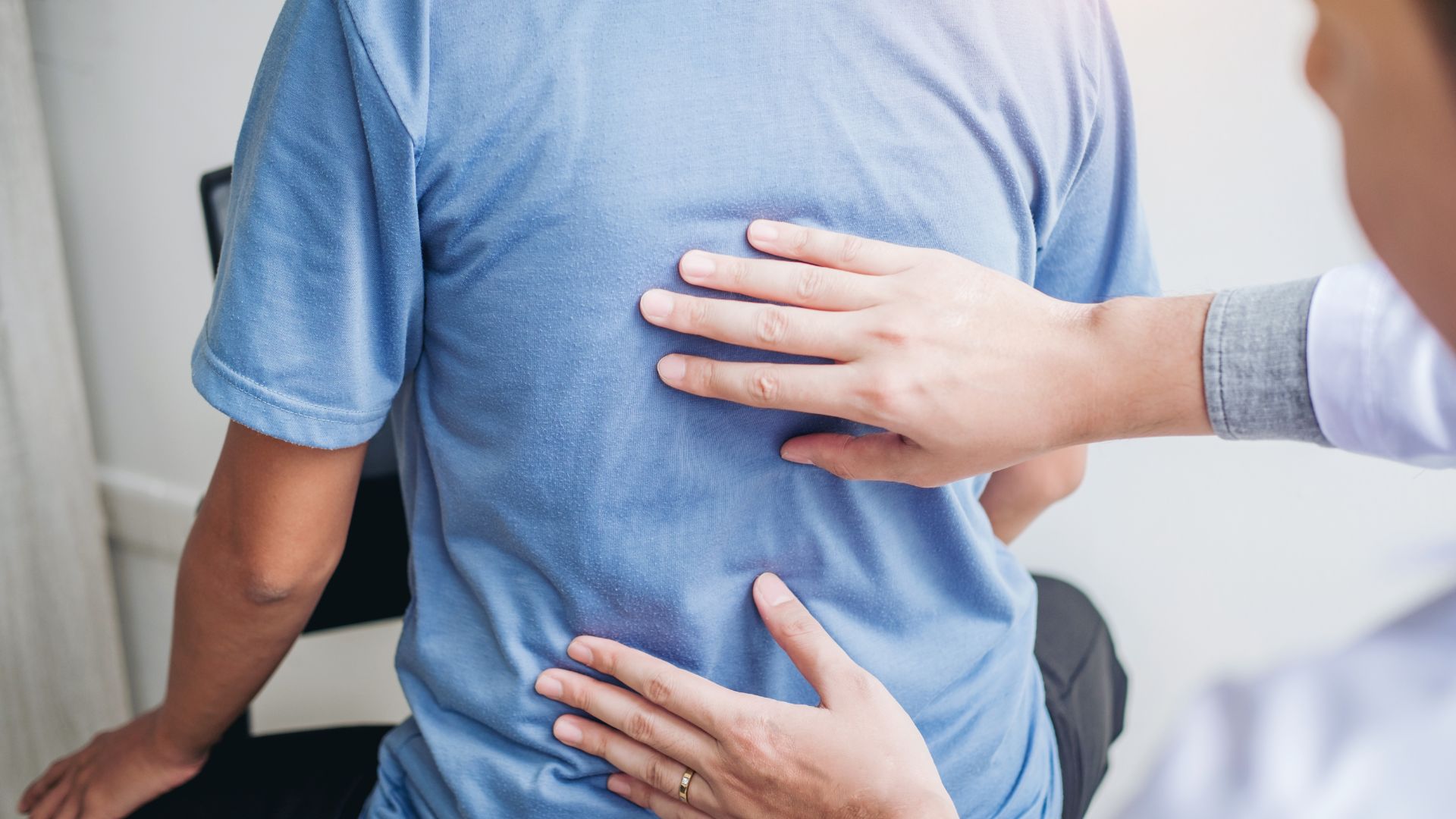 A healthcare professional in a white coat assesses a patient's back, placing hands on the upper and lower back. The patient wears a blue shirt.
