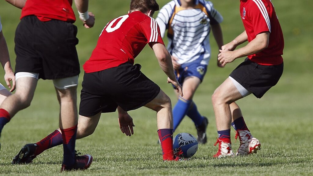 Three rugby players in red jerseys and black shorts are in a tackling stance, focusing on a player in blue and white, with a rugby ball on the grass.
