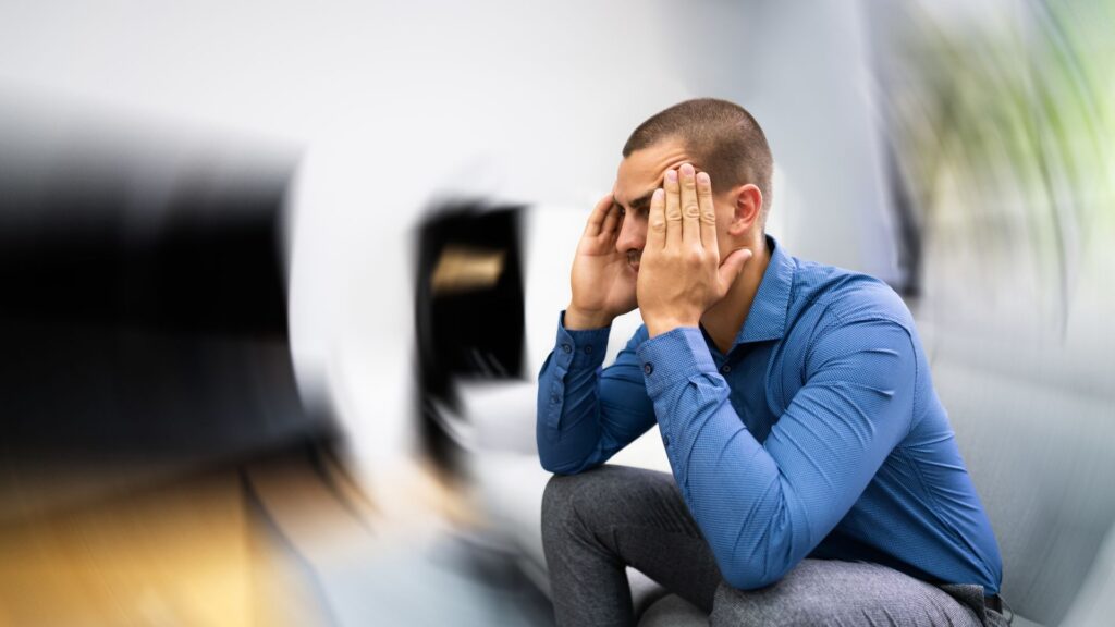 A man in a blue shirt sits, holding his head with both hands, conveying stress or dizziness. The background is blurred, emphasizing disorientation.