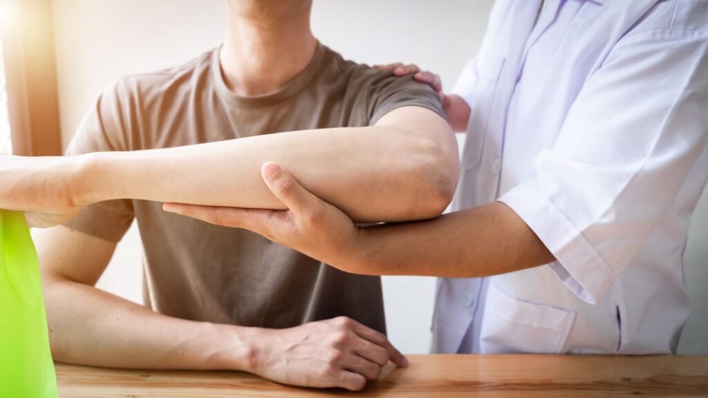 A therapist in a white coat supports a man's arm during a physical therapy session. The setting is bright and conveys a sense of care and rehabilitation.