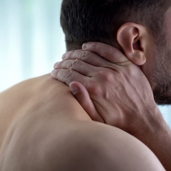 Man with bare shoulders and short hair holds his neck with both hands, suggesting discomfort or pain. Background is softly blurred, conveying a tense mood.